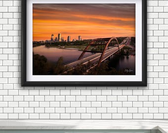 May include: A framed landscape photograph featuring a steel arch bridge spanning a river at sunset. The city skyline is visible in the distance under a vibrant orange and purple sky. The artwork is enclosed in a black frame with a white mat.