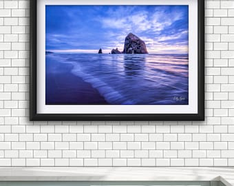 May include: A framed landscape photograph featuring a large sea stack rising from the ocean at twilight. The scene displays deep blue and purple hues across the sky and water, with gentle waves washing onto the sandy shore in the foreground.
