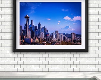 May include: A framed landscape photograph of the Seattle skyline featuring the Space Needle and a distant snow-capped mountain under a bright blue sky. The image is enclosed in a thick black frame with a white mat border, displaying urban architecture.