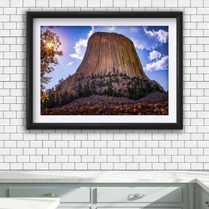 May include: A framed landscape photograph of Devils Tower, a tall igneous rock formation with vertical columns, surrounded by green trees and a rocky base under a bright blue sky with white clouds and a sun flare on the left side.