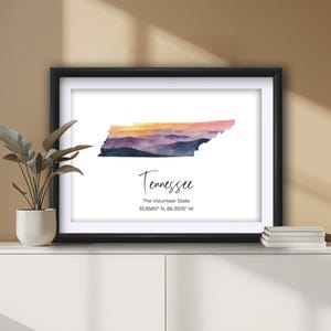 May include: Framed watercolor print of the state of Tennessee, featuring a colorful sunset over a mountain range. The print includes the state name and coordinates. The frame is black and the print is displayed on a white background.