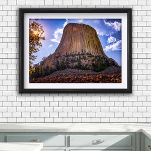 May include: Framed landscape photograph of Devils Tower National Monument. The image captures the iconic rock formation under a blue sky with scattered clouds. The sun shines through the trees on the left, illuminating the tower's textured surface and the surrounding landscape.