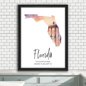 May include: A framed wall art print featuring a watercolor-style silhouette of the state of Florida filled with a sunset beach pier scene. Below the map, the text reads Florida, The Sunshine State, and the coordinates 28.6305 N, 82.4497 W.