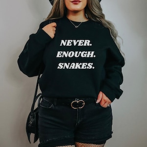 Può includere: Felpa nera con la scritta "NEVER. ENOUGH. SNAKES." a blocchi bianchi. La persona nell'immagine indossa pantaloncini neri, una cintura nera con fibbia dorata e una borsa nera.
