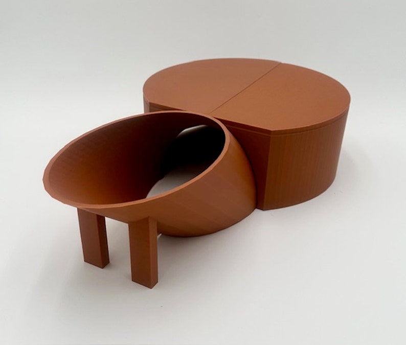 May include: A terracotta-colored ceramic object with a rounded top and a curved, hollow section with two legs. The object is made of two pieces that fit together.