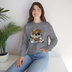 May include: A gray sweatshirt featuring a skull design with a snake wrapped around it, surrounded by white and gray flowers. The graphic is in shades of brown, black, and white. The sweatshirt has long sleeves and a crew neck.