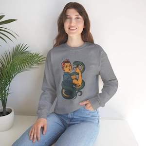 Peut inclure: Un sweat-shirt gris avec une illustration de style vintage d'une femme musclée avec un serpent enroulé autour d'elle. La femme porte un bandana rouge et une chemise bleue. Le serpent est vert et jaune.