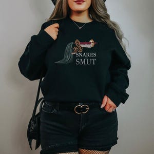 Könnte beinhalten: Schwarzes Sweatshirt mit dem weißen Schriftzug "Snakes and Smut". Die Grafik zeigt eine Schlange, die sich um ein Schwert windet. Das Sweatshirt ist ein lässiger Stil.