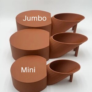 May include: Three terracotta-colored ceramic planters, labeled "Jumbo", "Mini", and "Mini". The planters are in the shape of a cylinder and a curved spout.