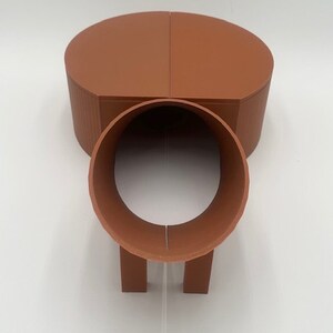 May include: A terracotta-colored, two-piece, 3D printed tunnel toy for small animals. The tunnel is made of two half-circles that connect to form a tube. The tunnel has two short legs on the bottom.