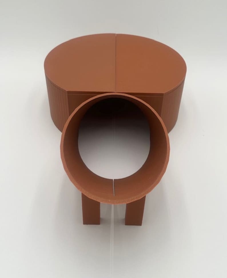 May include: A terracotta-colored, two-piece, 3D printed tunnel toy for small animals. The tunnel is made of two half-circles that connect to form a tube. The tunnel has two short legs on the bottom.