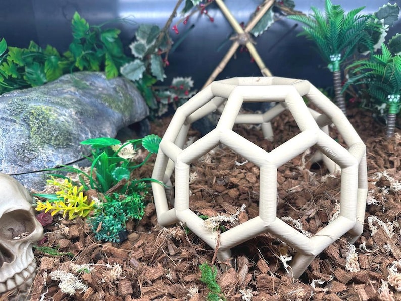 May include: A white geodesic dome made of plastic, sitting on a bed of brown wood chips. The dome is a reptile hide for a terrarium.