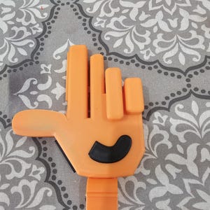 May include: A 3D-printed orange hand with a black curved shape on the palm. The hand is in a gesture of holding something. The fingers are straight and the thumb is extended. The hand is attached to a rectangular base.