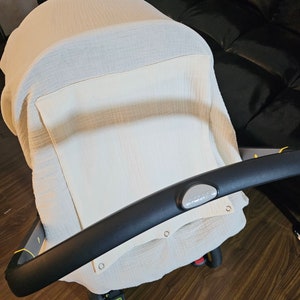 May include: A white muslin stroller cover with a white fabric panel and black stroller handle. The cover has small holes for ventilation.