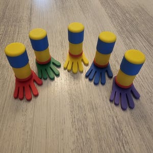 May include: Five colorful hand-shaped bottle stoppers. Each stopper has a yellow top, blue band, red band, and a hand in a different color: red, green, yellow, blue, and purple. These decorative stoppers add a playful touch to any bottle.