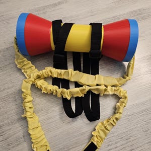 Puede incluir: Un dispositivo de ejercicio colorido con un centro amarillo y extremos rojos, cada uno con un borde azul. Se adjuntan correas negras y bandas elásticas amarillas con cierres de velcro, lo que sugiere una herramienta de fitness o rehabilitación.