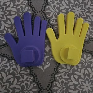 May include: Two hand-shaped objects, one purple and one yellow, each with a square base. The hands are positioned on a patterned gray surface. The hands are made of a solid material.