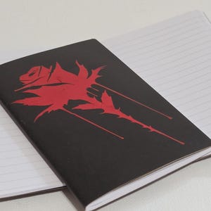 Puede incluir: Un cuaderno negro con un diseño de rosa roja y páginas rayadas. La ilustración de la rosa está estilizada y cubre parte de la portada. El cuaderno está abierto, revelando las páginas interiores blancas y rayadas.
