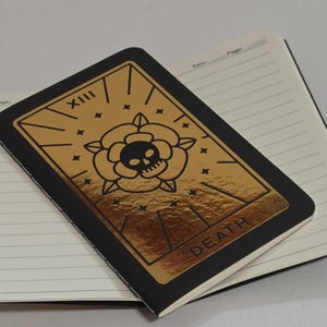 Puede incluir: Un cuaderno negro con páginas rayadas, cubierto por un cuaderno más pequeño con un diseño de lámina dorada. El diseño dorado incluye una calavera, una rosa y la palabra "DEATH". El cuaderno superior tiene el número romano "XIII".