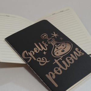 Puede incluir: Cuaderno negro con las palabras "Spells & Potions" en lámina de color cobre. El cuaderno tiene páginas rayadas y un diseño de botella de poción con una calavera y estrellas. El cuaderno está abierto, revelando las páginas interiores.