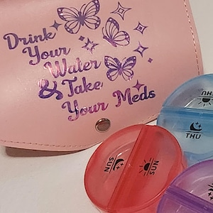 May include: A pink pill case with the text "Drink Your Water & Take Your Meds" and butterfly graphics. Several colorful, oval-shaped pill organizers are in the foreground, each labeled with a day of the week.