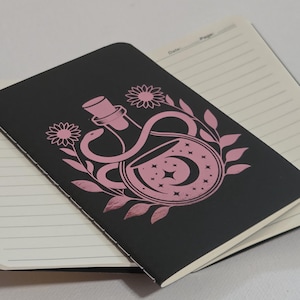 Puede incluir: Un cuaderno negro con una ilustración rosa de una botella de poción, una serpiente, una luna y estrellas. El cuaderno está abierto, revelando páginas rayadas. El diseño está rodeado de flores y hojas.