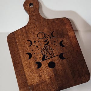 May include: A wooden chopping board with a handle, featuring a decorative design. The design includes a house, crescent moons, stars, and bats. The board is brown with a smooth finish.