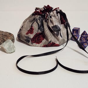 Pode incluir: Um saco de dados de tecido com um padrão floral de rosas vermelhas, um cordão preto e um conjunto de dados roxos. Várias pedras preciosas também estão presentes. O saco foi concebido para guardar dados.
