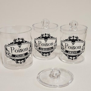 May include: Three clear apothecary jars with lids, labeled "Poison" in black script with skull and crossbones emblems. The jars are labeled "Cyanide", "Strychnine", and "Arsenic". One lid is off, revealing the contents.