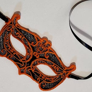 May include: An orange and black lace masquerade mask with a black satin ribbon tie. The mask features an intricate floral design and is suitable for a costume party or masquerade ball.