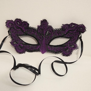 May include: A masquerade mask crafted from black and purple lace with an intricate embroidered floral pattern. The mask features two eye openings and is secured with thin black satin ribbons attached to each side for tying around the head.