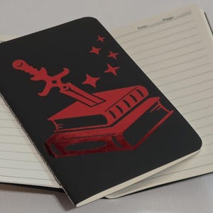 Puede incluir: Un cuaderno negro con un diseño rojo de una espada que atraviesa dos libros y estrellas. El cuaderno está abierto, revelando páginas blancas rayadas. El cuaderno está sobre una superficie blanca.