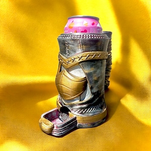 May include: A decorative can cooler in a warrior design, featuring a gold and silver color scheme. The cooler has a mask-like front and a small tray at the base. A pink can is inside the cooler.