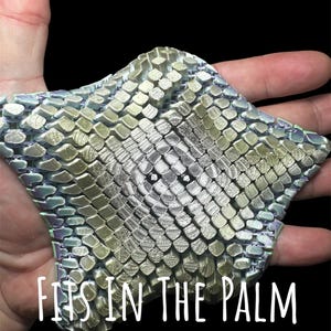 May include: A textured, star-shaped object with a metallic, scale-like pattern in shades of silver, green, and gold. The text "FITS IN THE PALM OF YOUR HAND" is displayed in white. The object is held in an open hand against a black background.