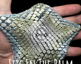 May include: A textured, star-shaped object with a metallic, scale-like pattern in shades of silver, green, and gold. The text "FITS IN THE PALM OF YOUR HAND" is displayed in white. The object is held in an open hand against a black background.