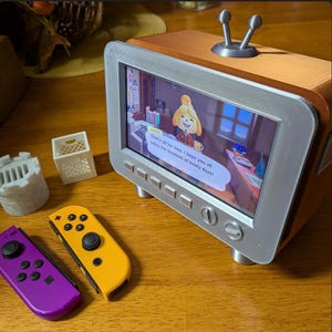 May include: A wooden-look TV with a silver frame displays a video game scene. The screen shows a character with text that reads, "That's all for now, I hope you all enjoy the loveliest of lovely days!" A purple and yellow game controller and miniature accessories are nearby.