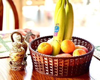 May include: A wicker basket filled with yellow bananas and orange citrus fruit. The basket is on a wooden table with a patterned placemat.