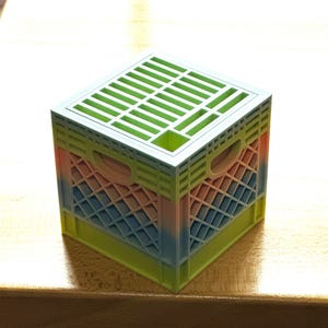 May include: A small, pastel-colored plastic crate with a green top and a grid pattern. The crate is made of a translucent plastic and has a rainbow ombre effect.