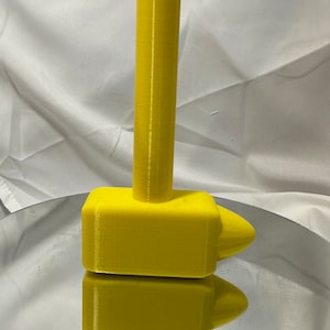 May include: A bright yellow hammer-shaped object with a cylindrical handle and a rectangular head with a conical end. The object is 3D-printed and sits on a reflective surface, with its reflection visible below.
