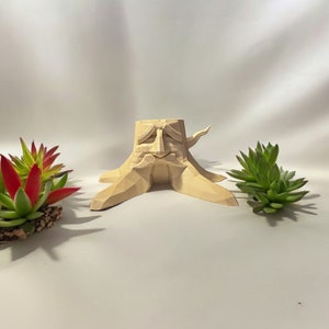 May include: A light brown, 3D printed, geometric tree stump planter with a face carved into the front. The planter is sitting on a white surface with two small green succulent plants.