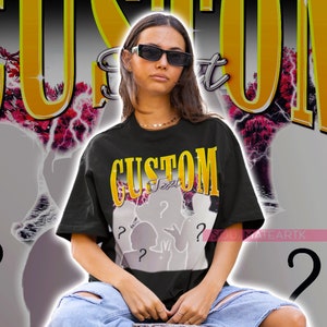May include: Black t-shirt with the word "CUSTOM" in gold and yellow lettering. The shirt features a graphic with silhouettes and question marks. The model is wearing sunglasses and ripped blue jeans.