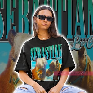 May include: A woman wearing a black t-shirt with a graphic of two miniature horses and the text "SERASTIAN Lil'" in blue and green. She is wearing blue jeans with rips in the knees and sunglasses.