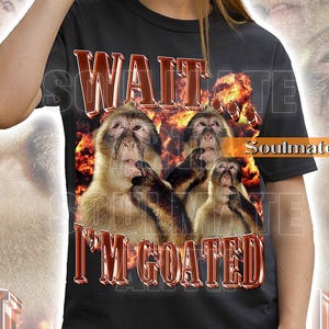 Peut inclure: T-shirt noir avec l'inscription "WAIT I'M GOATED" en rouge métallisé. Le t-shirt présente un graphisme de singes avec un fond enflammé. La personne sur l'image porte un bob noir et des lunettes de soleil.