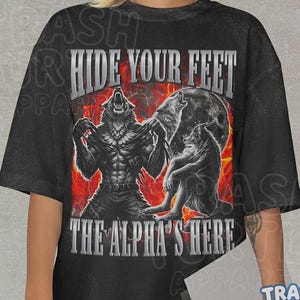 Puede incluir: Camiseta negra con un gráfico de un hombre lobo, una luna llena y el texto "HIDE YOUR FEET THE ALPHA'S HERE." El diseño presenta un hombre lobo rasgando su camisa, con un fondo rojo y naranja llameante. La camiseta es de Trash Turtle Apparel.