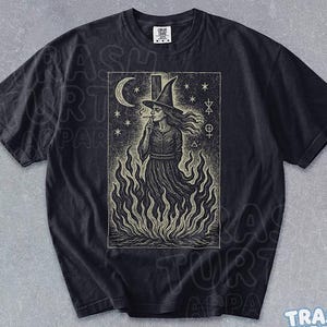 May include: Black t-shirt with a detailed illustration of a witch in a tall hat, smoking, and standing in flames. The design includes a crescent moon, stars, and mystical symbols. The text "BLACK" is at the bottom.