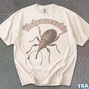 May include: A natural ivory short-sleeve t-shirt featuring a brown, detailed illustration of a weevil insect centered on the chest. Above the insect, the text "Unbeweevable" is printed in a curved, brown, retro-style font on the fabric.