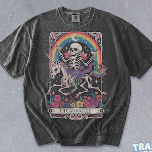 May include: A charcoal gray short-sleeve t-shirt featuring a graphic of a skeleton riding a unicorn under a rainbow. The design includes flowers, stars, and the text THE AUDACITY. The shirt has a crew neckline and a vintage-style washed finish.