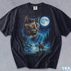 Puede incluir: Una camiseta negra de manga corta con un estampado gráfico de la cara de un gato grande mirando a un gato más pequeño de pie sobre una roca junto a una cascada en un bosque por la noche bajo una luna llena y un cielo estrellado.