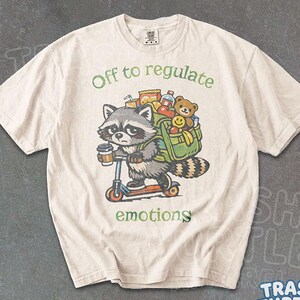 Puede incluir: Una camiseta de color marfil con el gráfico de un mapache en un patinete, llevando una mochila verde llena de aperitivos, un osito de peluche y una bebida. El mapache sostiene un café. El texto dice Off to regulate emotions.