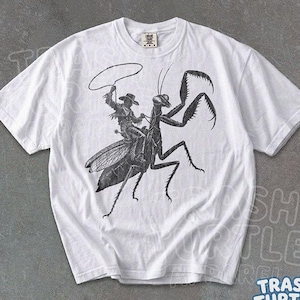 Könnte beinhalten: Ein weißes Kurzarm-T-Shirt mit einer schwarzen Grafik im Tintenstil, die einen Cowboy zeigt, der auf einer großen Gottesanbeterin reitet und ein Lasso schwingt. Das Shirt hat einen Rundhalsausschnitt und ein Etikett mit Comfort Colors.
